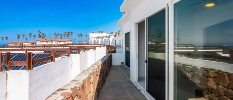 Un balcone con un muro in pietra, una ringhiera in legno e vista su palme e edifici.