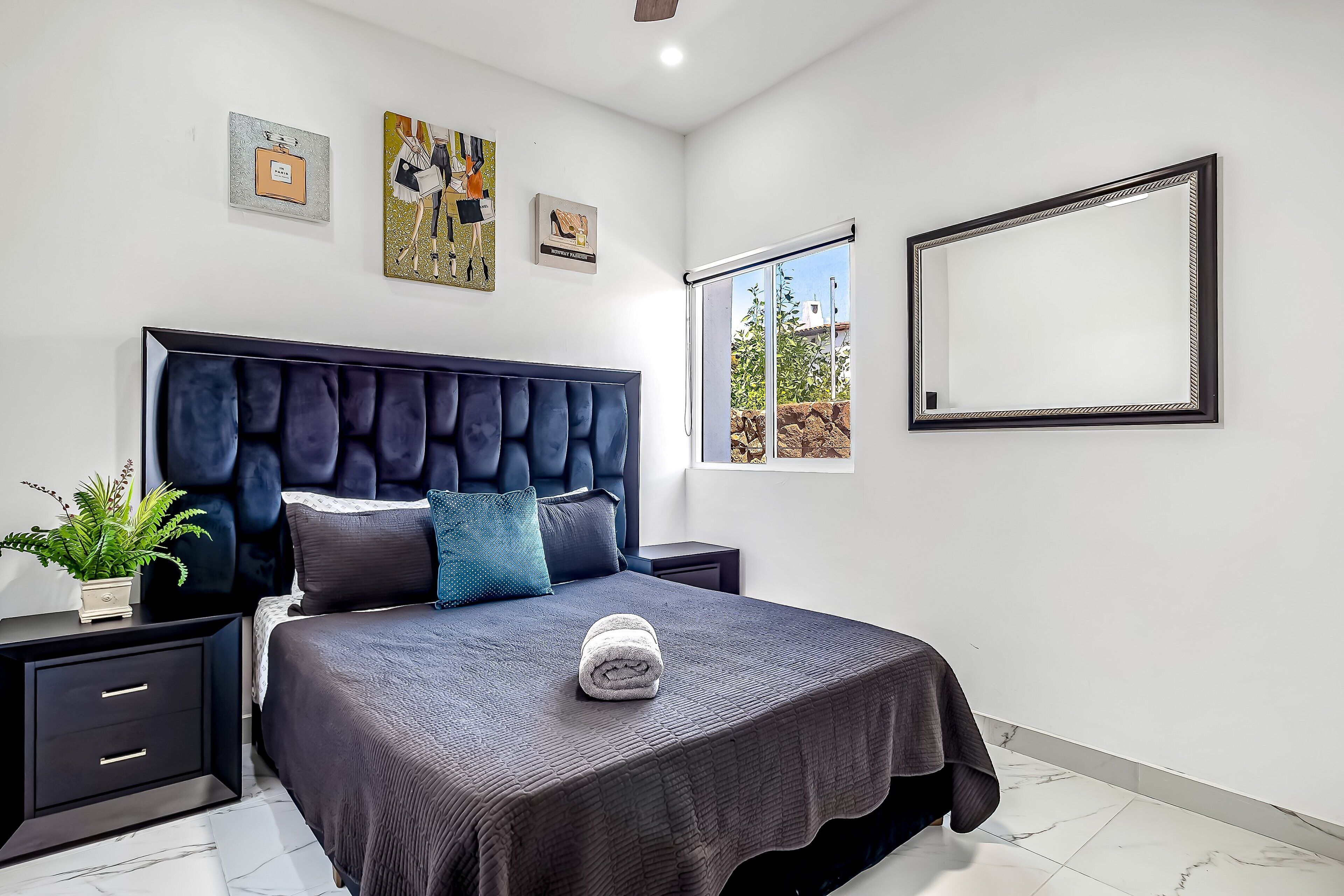 A modern bedroom with a large bed, a headboard, a nightstand, a potted plant, and artwork on the wall.