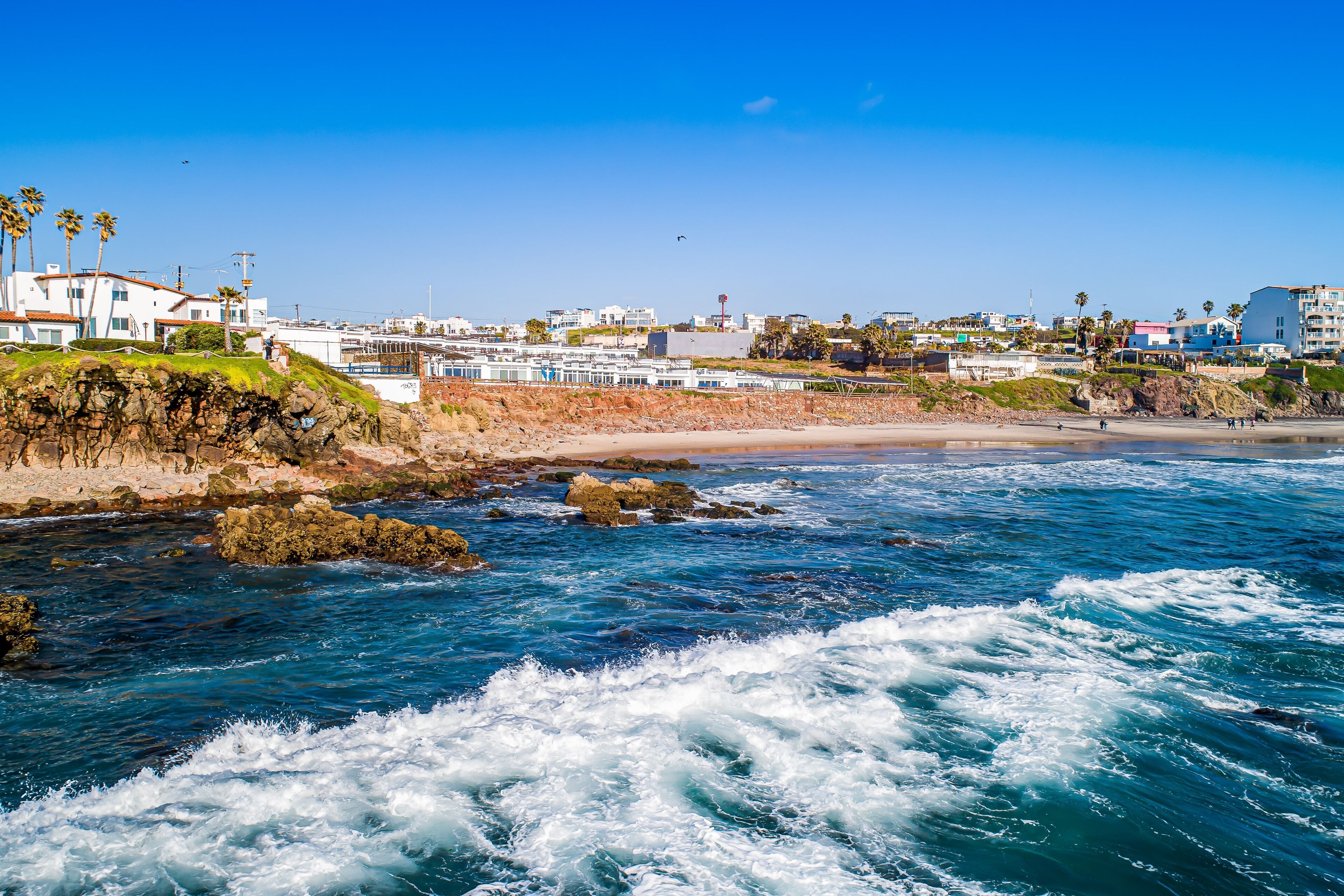 A coastal view with rocky shoreline, waves, and buildings in the background.