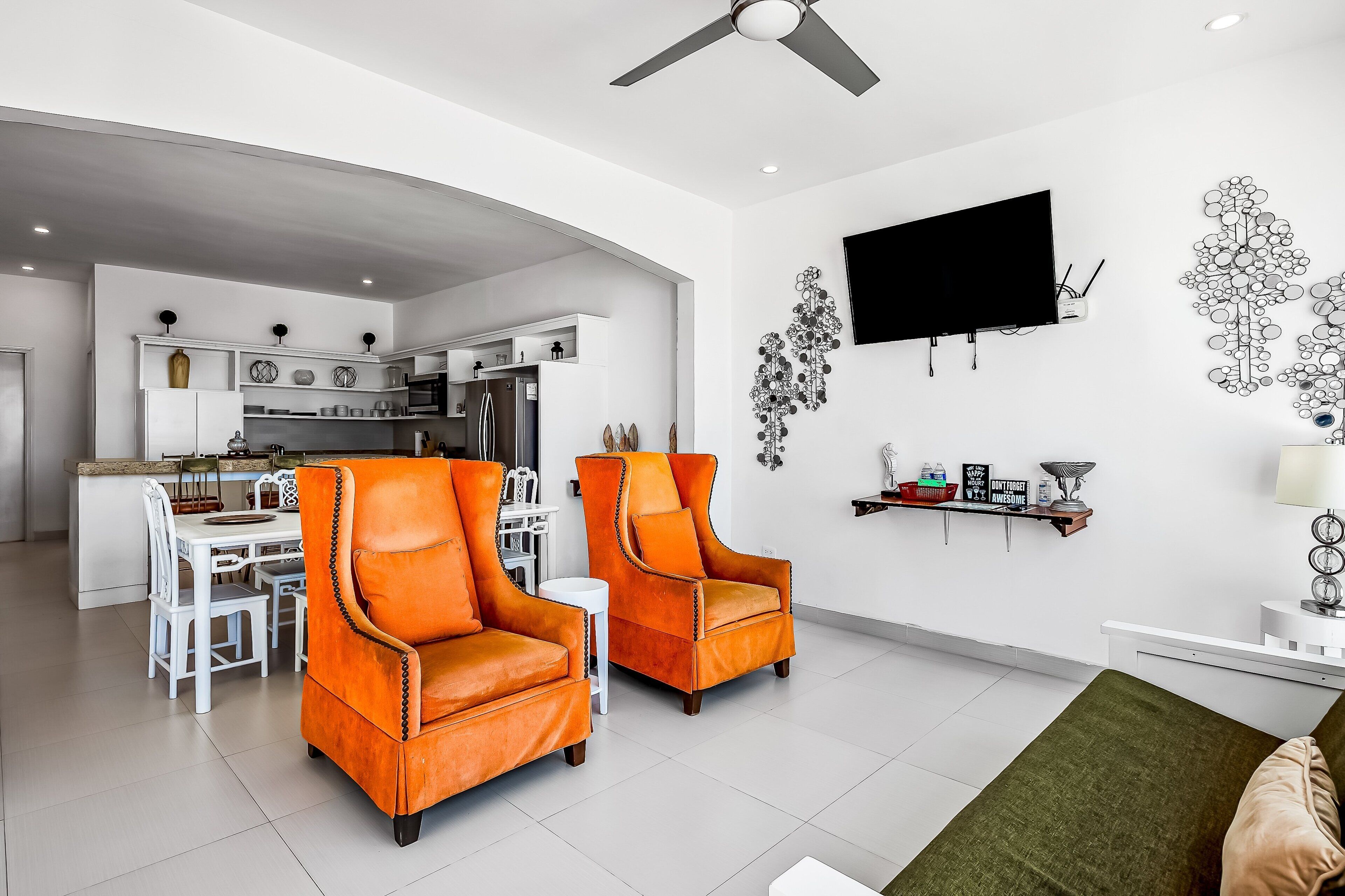 A modern living room with two orange armchairs, a dining area, a kitchen, and a flat-screen TV mounted on the wall.
