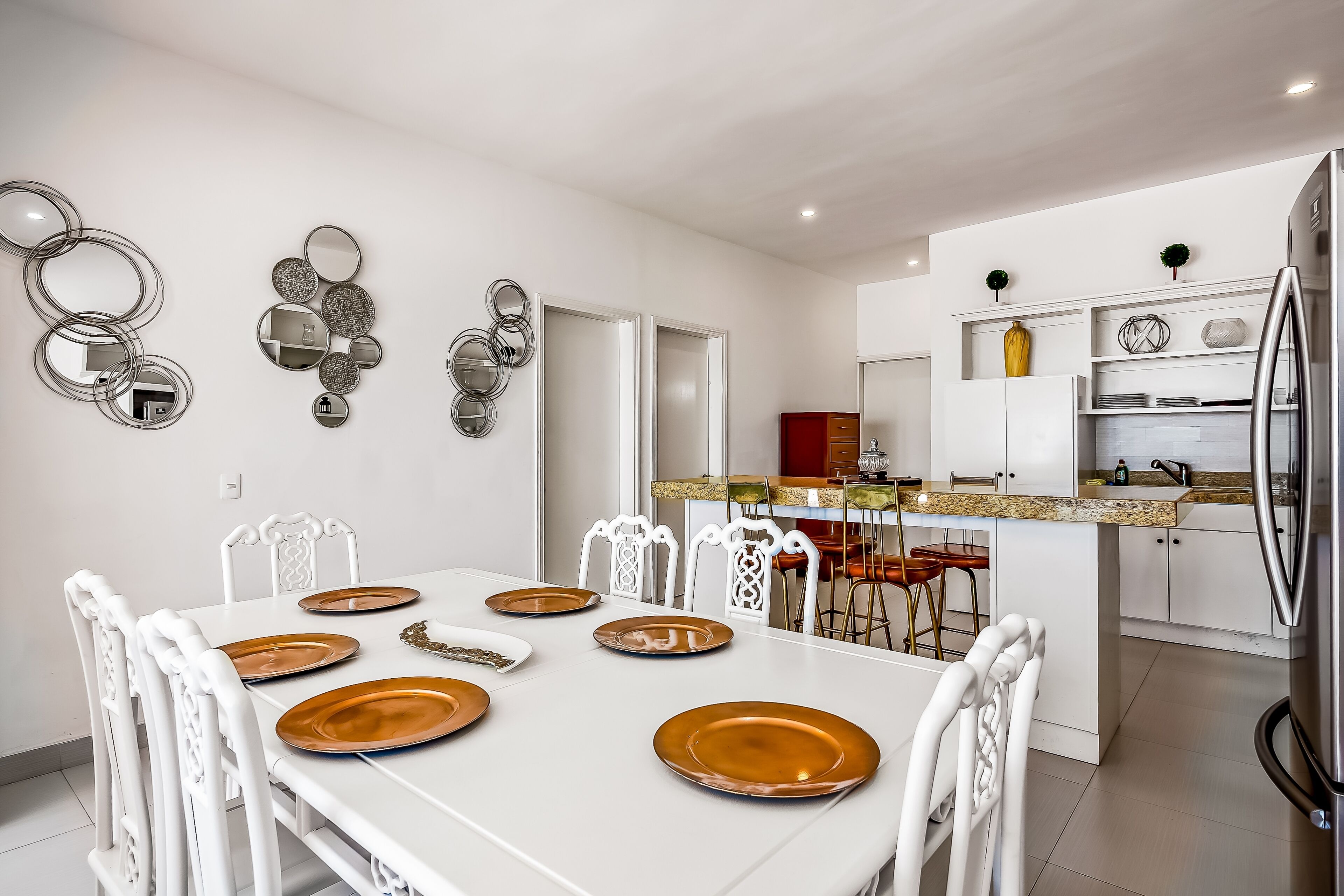 Cucina moderna con mobili bianchi, frigorifero in acciaio inossidabile e zona pranzo con sedie bianche e piatti marroni.