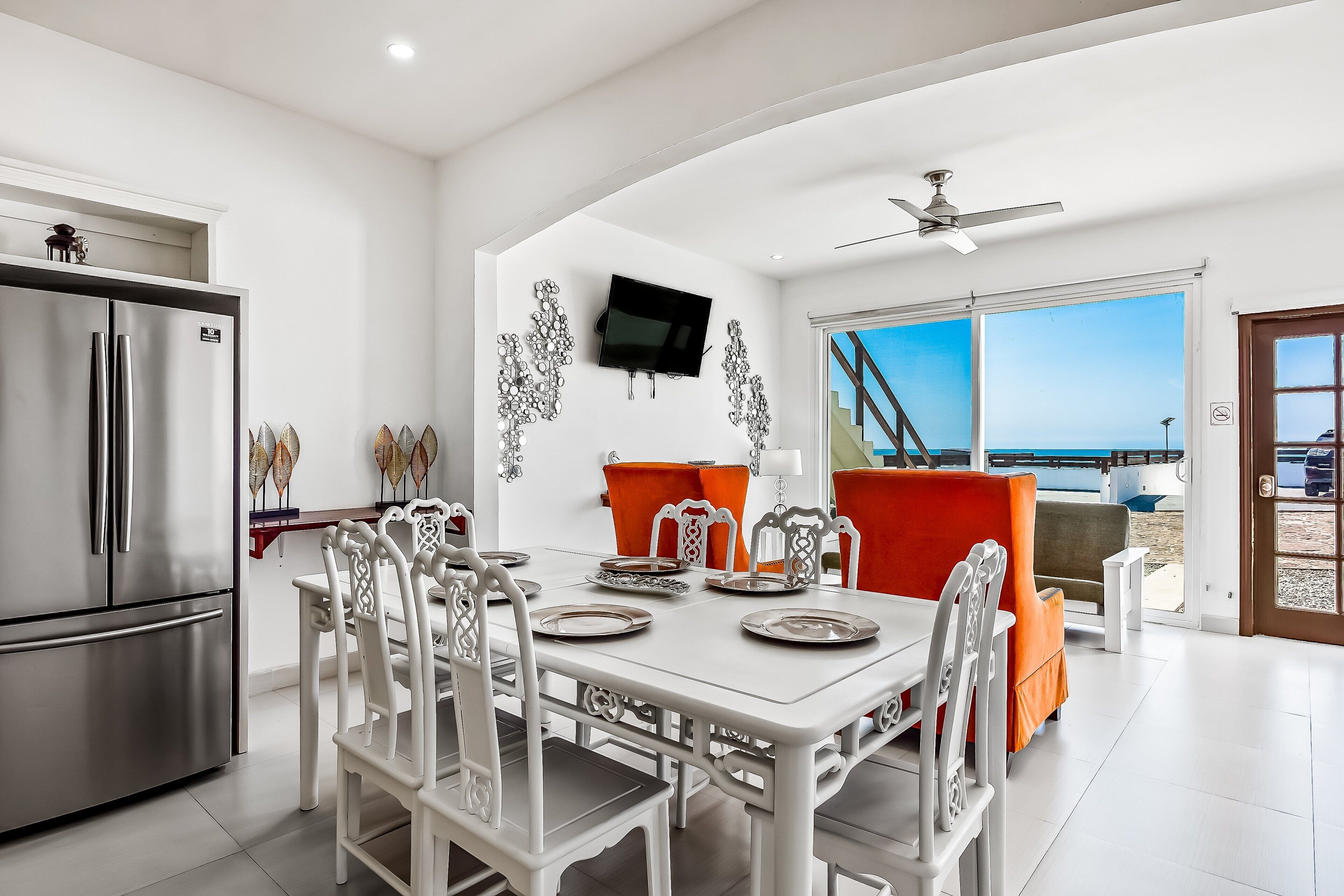 A modern kitchen with a stainless steel refrigerator, a dining area with white chairs and a table set for four, and a large window offering a view of the beach.