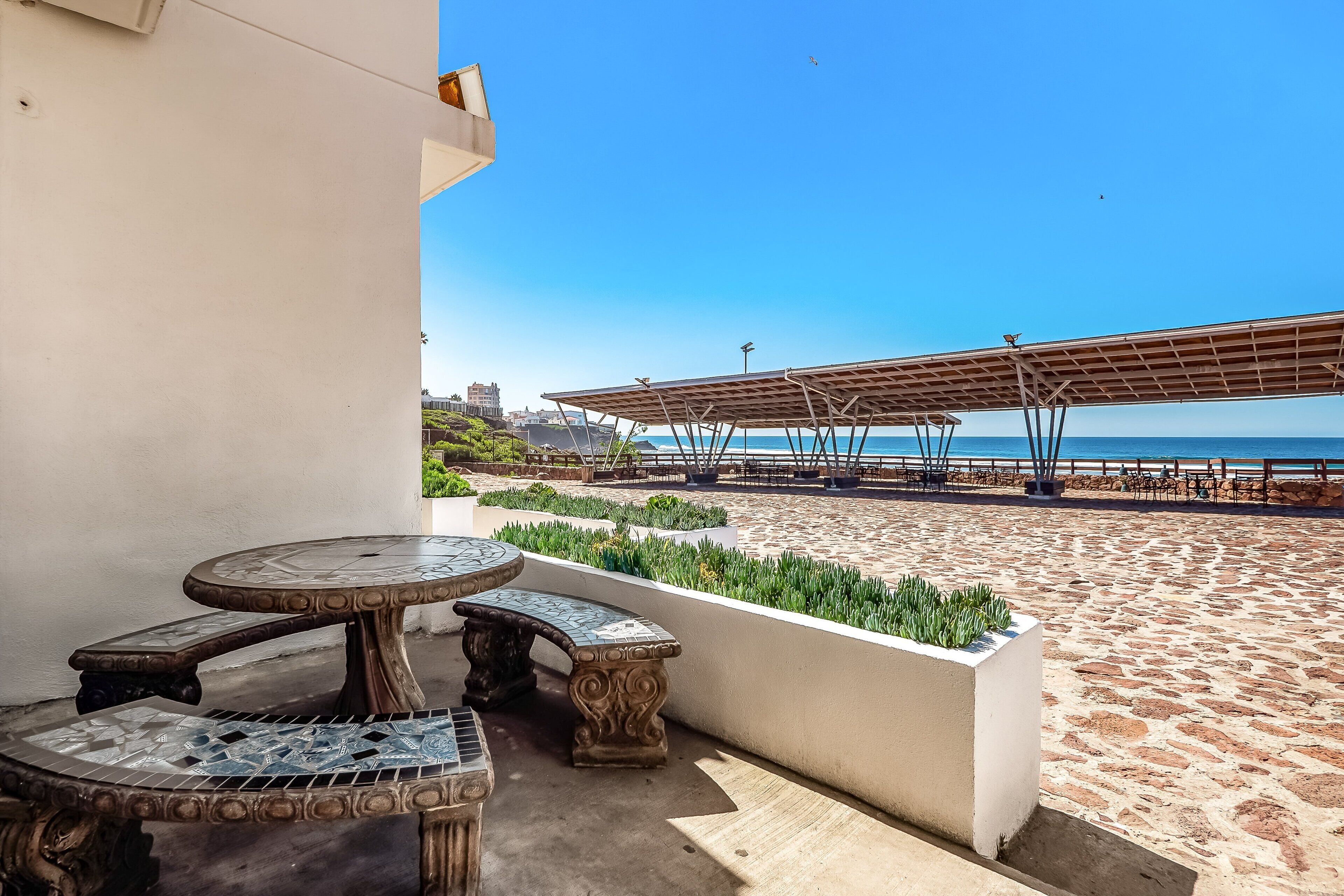 A patio with a round table and benches, a covered area with a view of the beach, and a white wall.