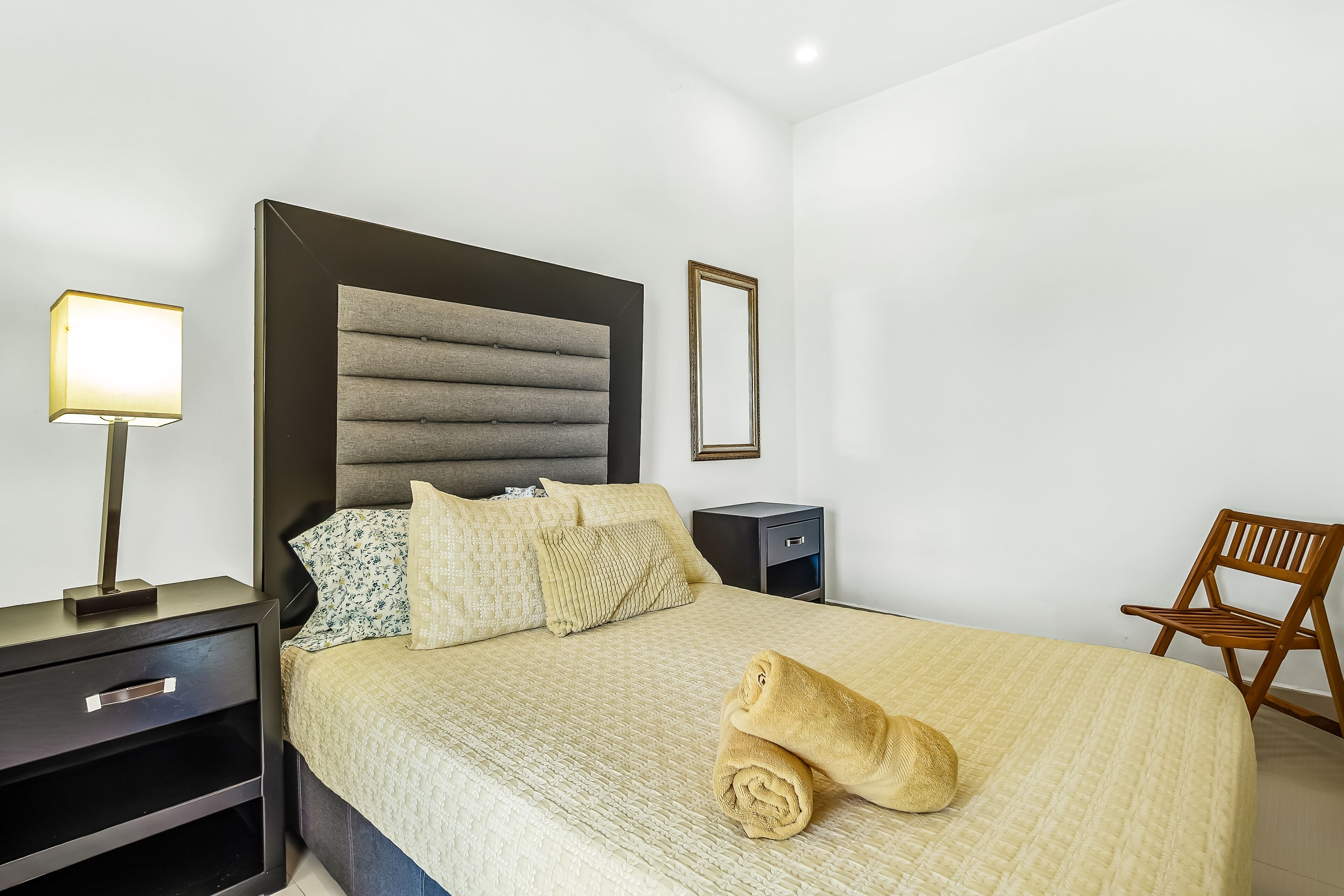 A neatly made bed with a rolled towel, a bedside lamp, a small nightstand, and a wooden chair.