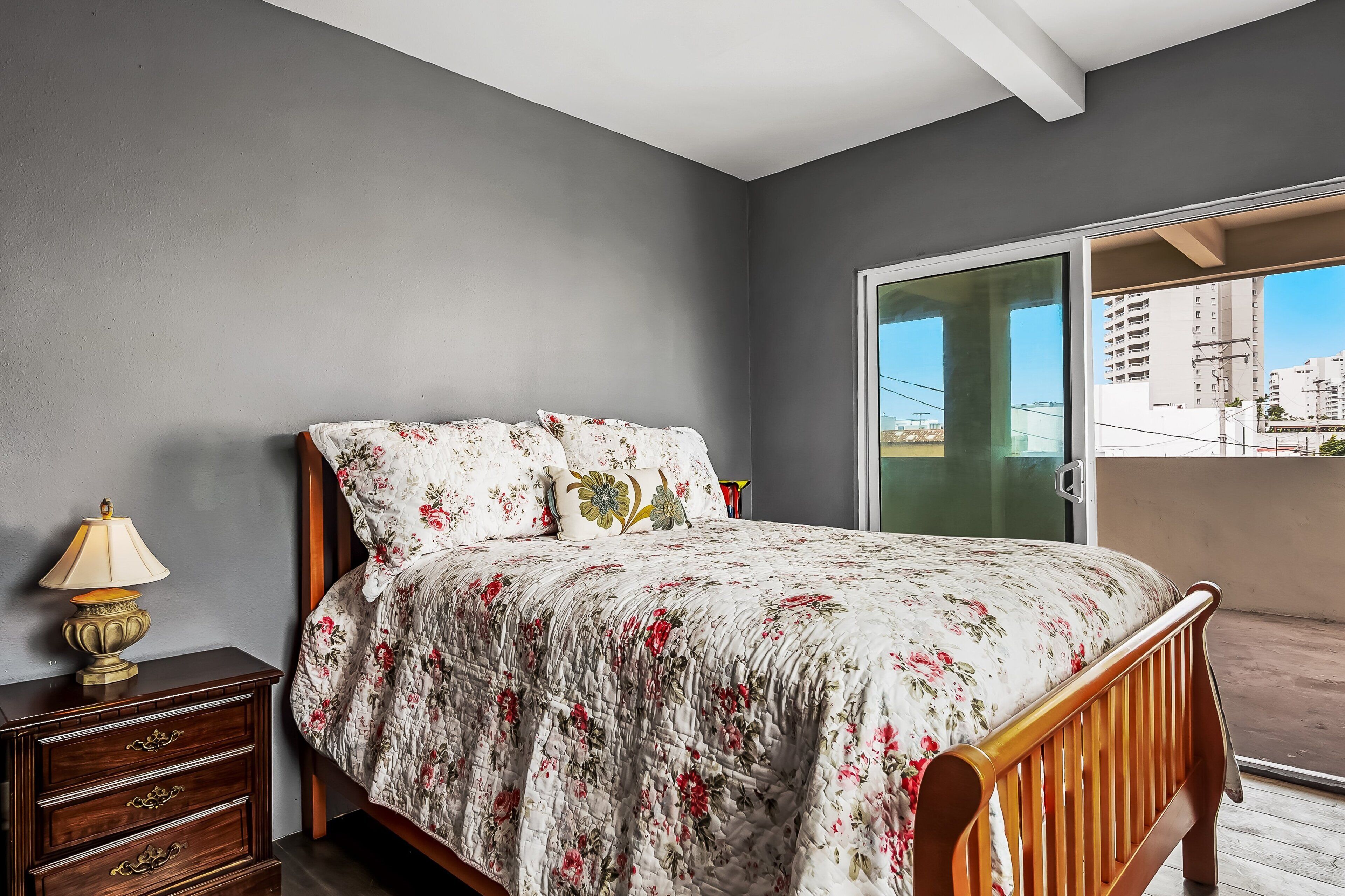 A bedroom with a bed, a nightstand, a lamp, and a view of buildings through a glass door.