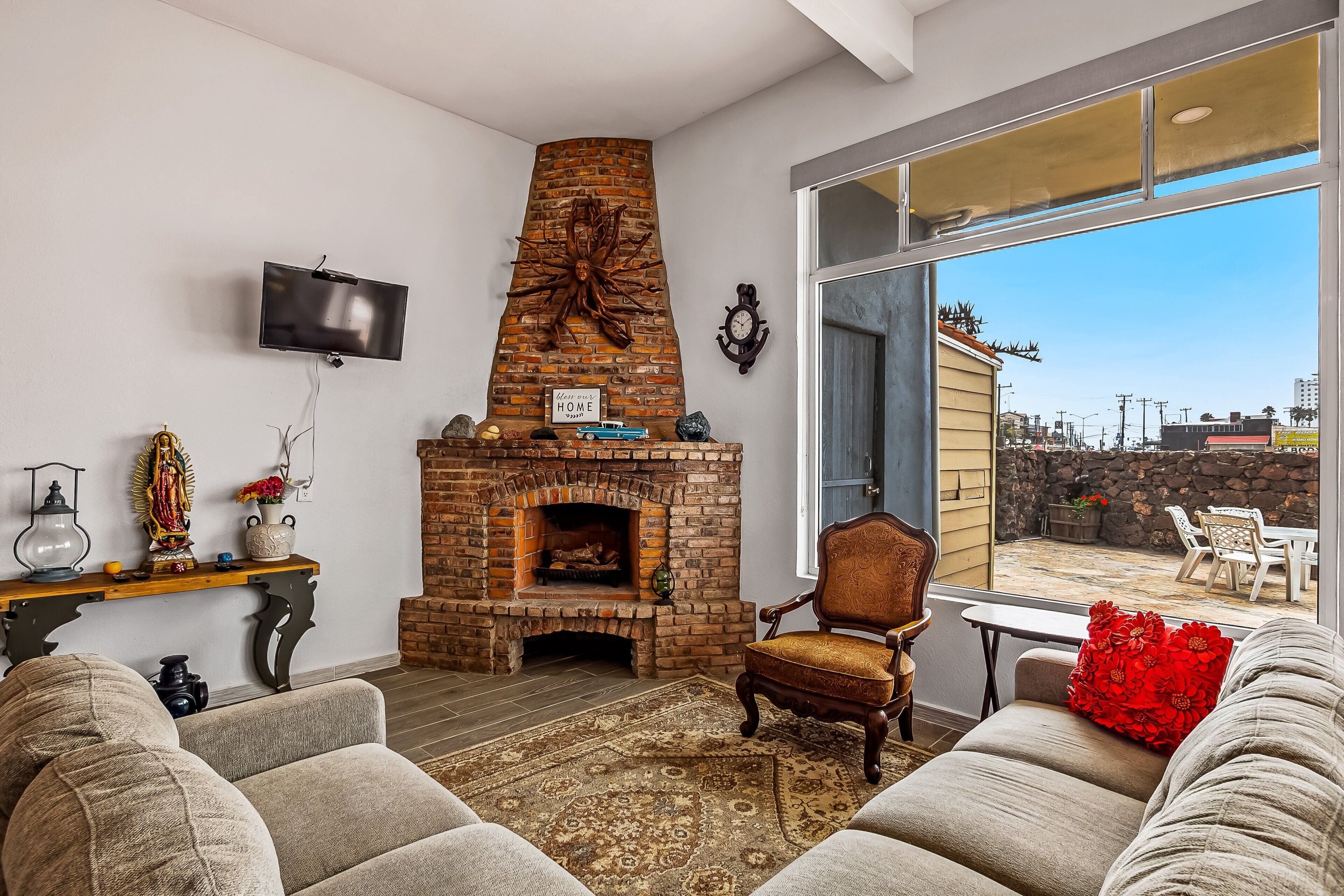 A living room with a brick fireplace, a television mounted on the wall, and a large window overlooking a patio area.