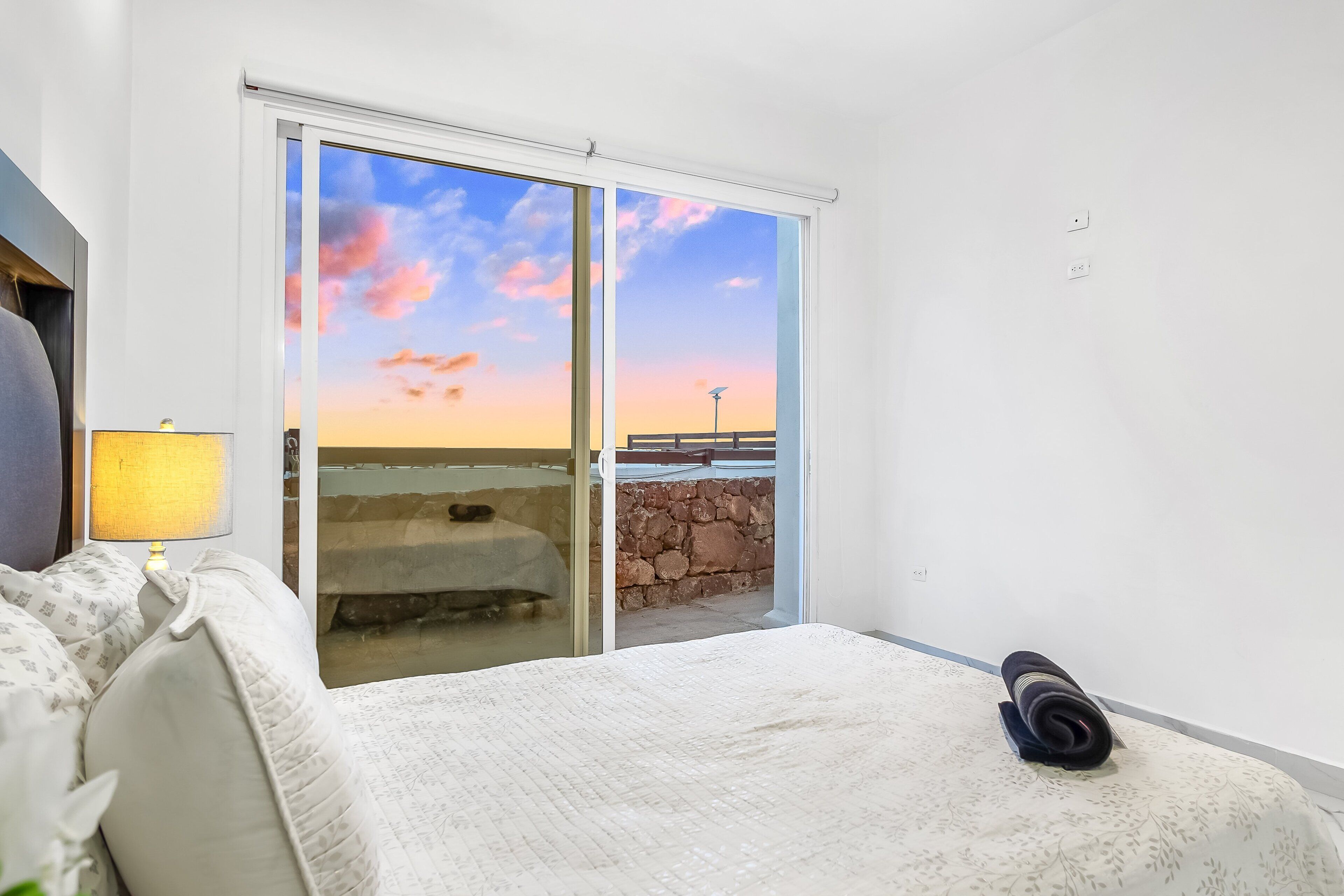 A bedroom with a bed, a lamp, a view of the sunset, and a balcony.