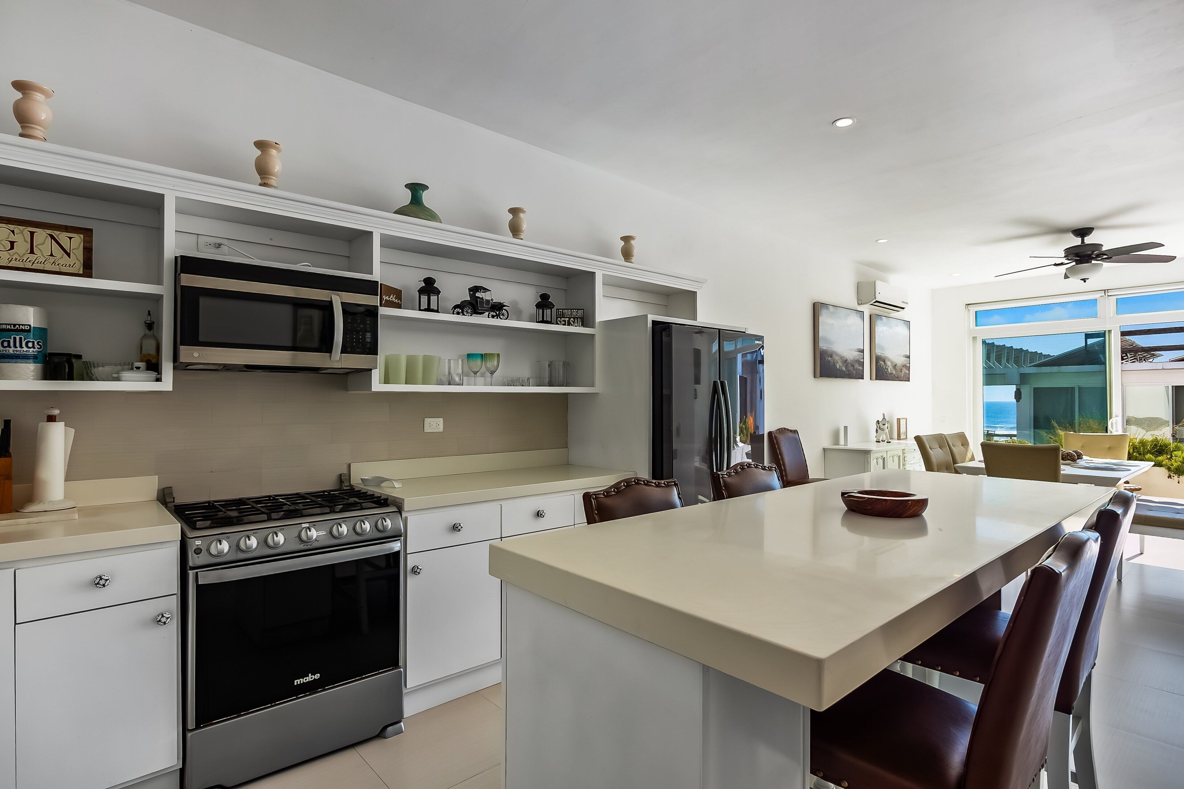 A modern kitchen with white cabinetry, a stove, microwave, and refrigerator. There is a dining area with a large island and chairs.