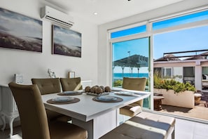 A dining area with a white table, beige chairs, and a view of the ocean through large windows. - Peaceful Dog-friendly Villa With Outdoor Pool, Rooftop Patio, Beach Views, & AC (Playas de Rosarito)