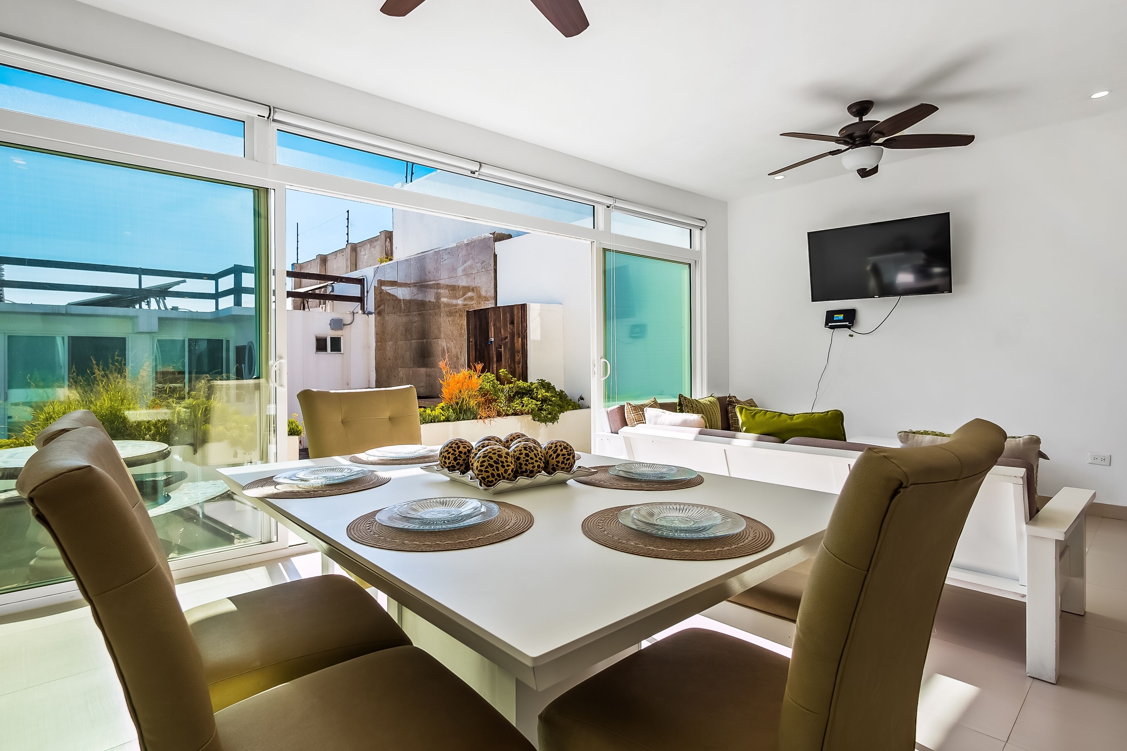 A modern dining area with a glass table, beige chairs, and a ceiling fan.
