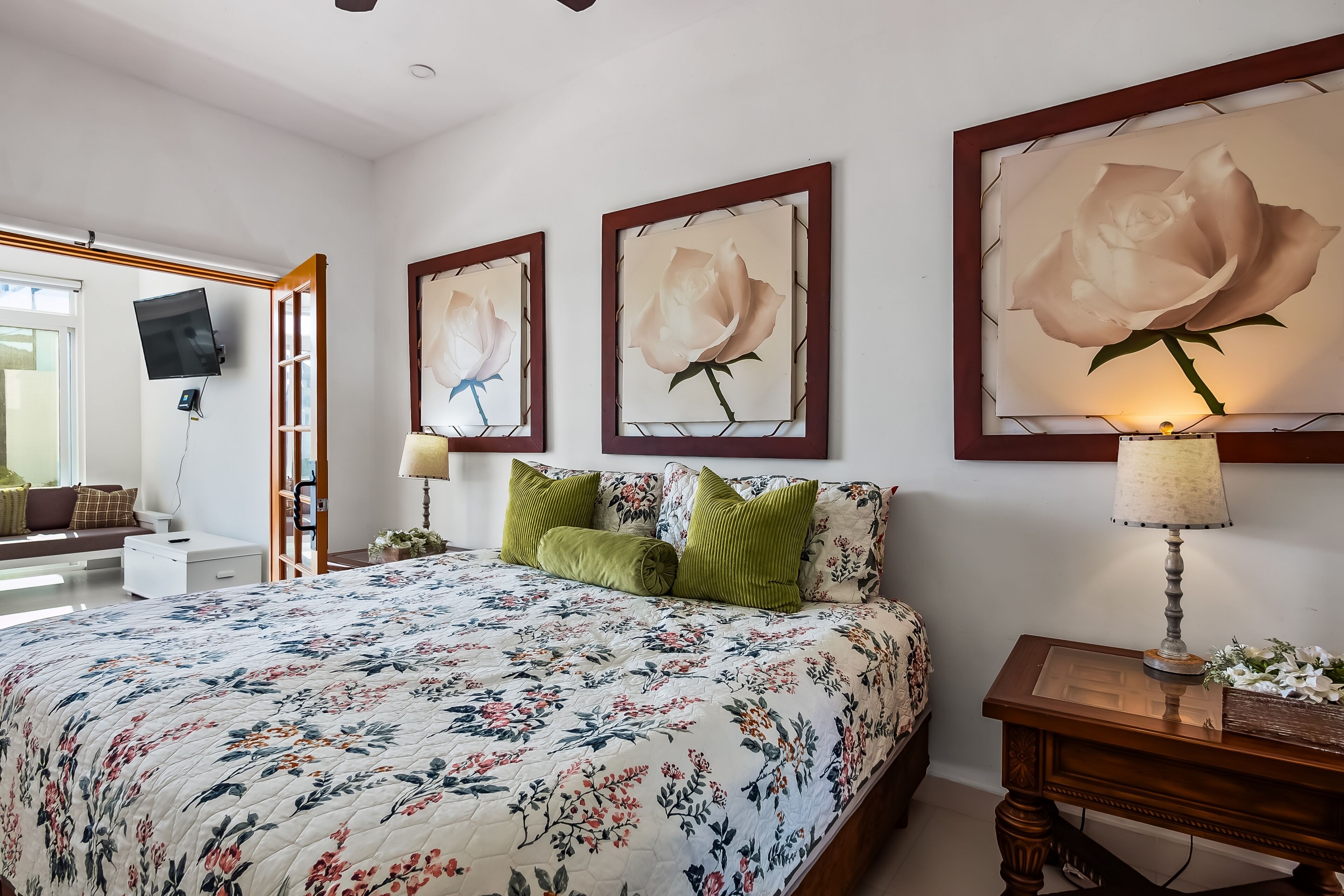 A bedroom with a floral bedspread, a wooden bedside table with a lamp, and three framed floral paintings on the wall.
