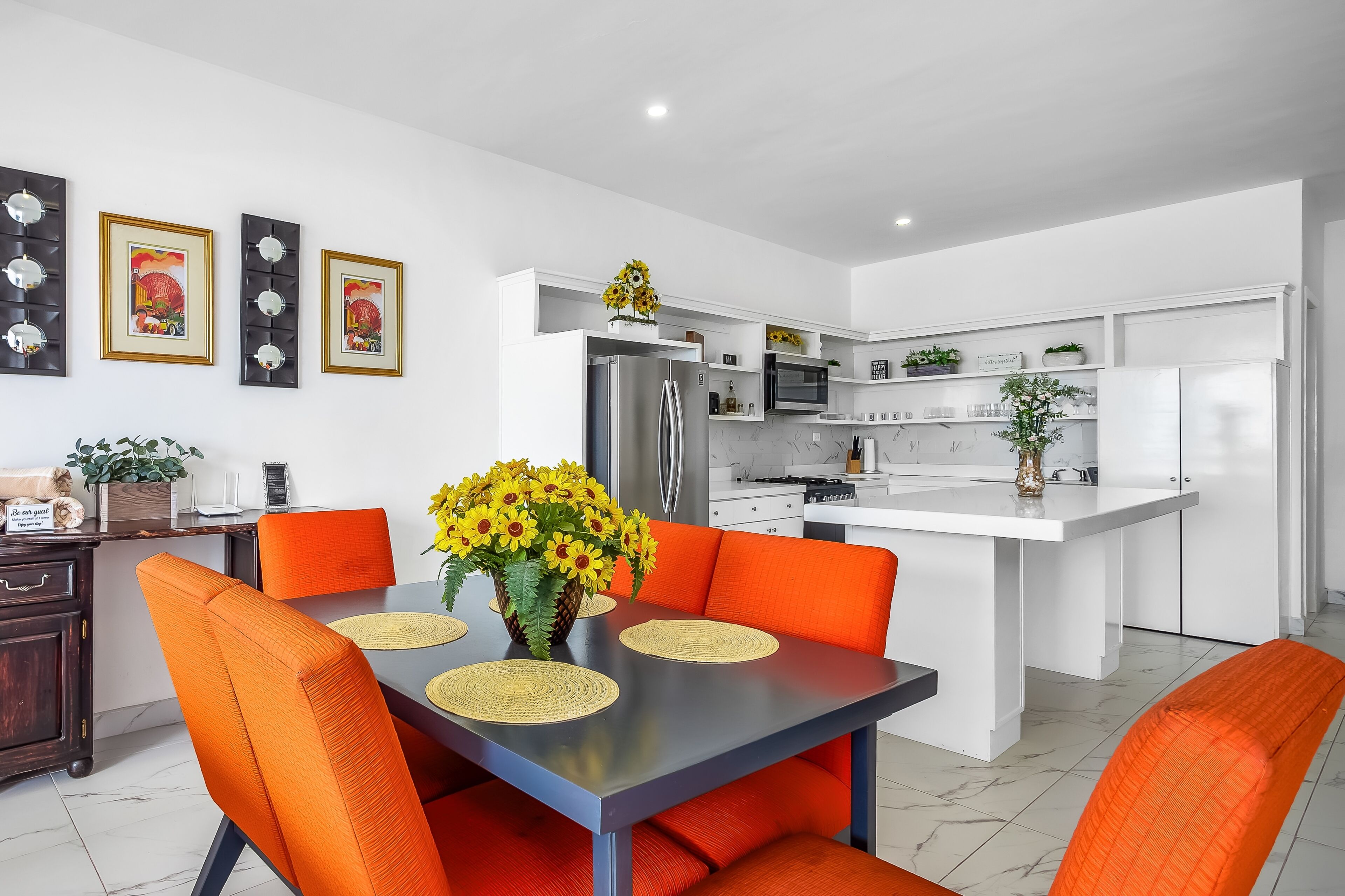 A modern kitchen with white cabinetry, a central island, and a dining area with orange chairs and a table.