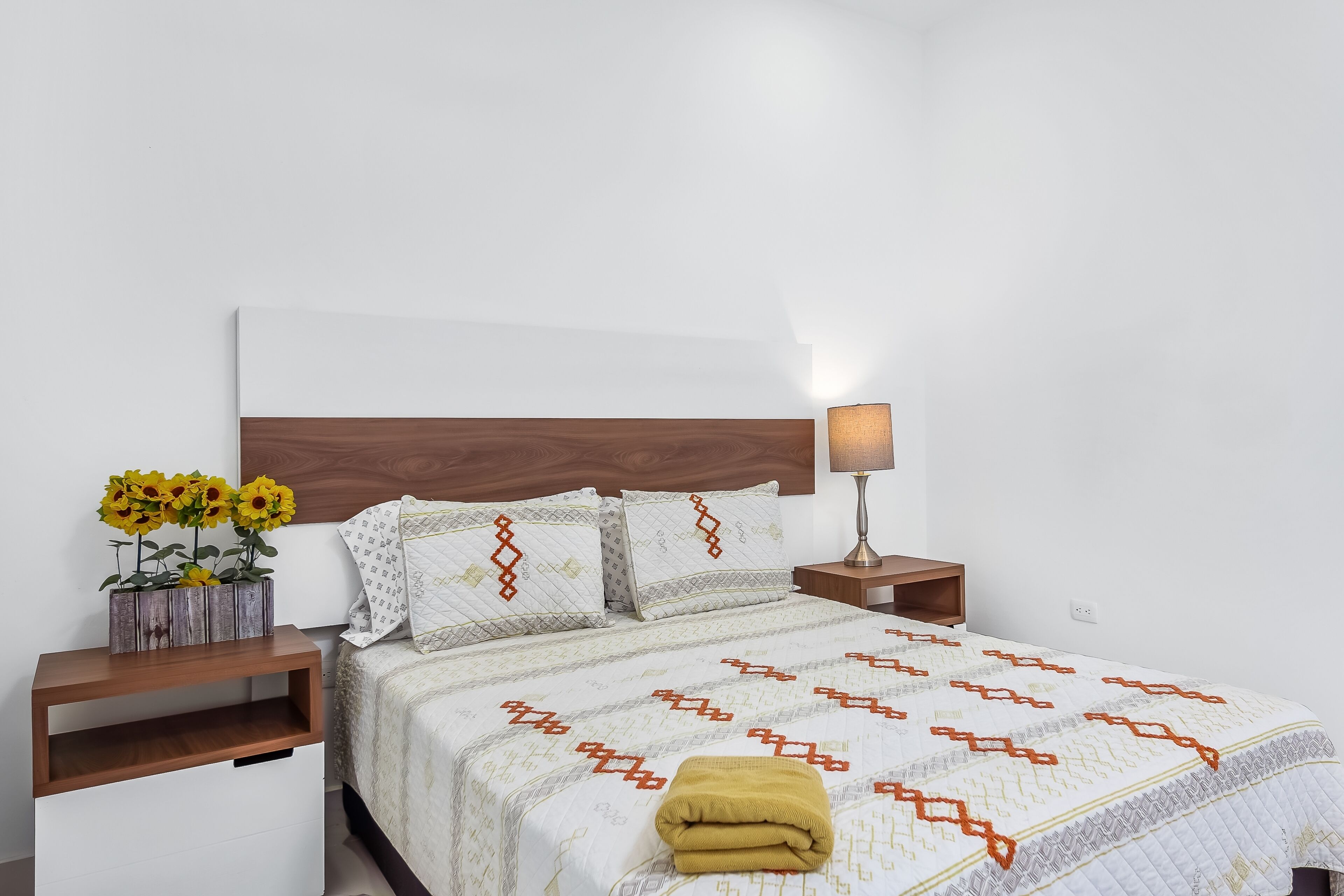 A neatly made bed with a patterned bedspread, a bedside lamp, and a wooden nightstand with a vase of sunflowers.