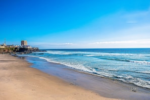 A beach with a building on the left, waves, and a clear sky. - Dog-friendly Home With Partial Ocean View, Heated Pool, Rooftop Terrace (Playas de Rosarito)