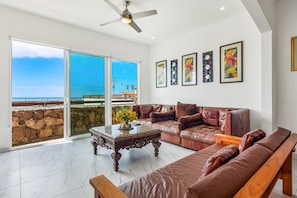 A spacious living room with a large sofa, a coffee table, and a view of the ocean through a sliding glass door. - Dog-friendly Home With Partial Ocean View, Heated Pool, Rooftop Terrace (Playas de Rosarito)