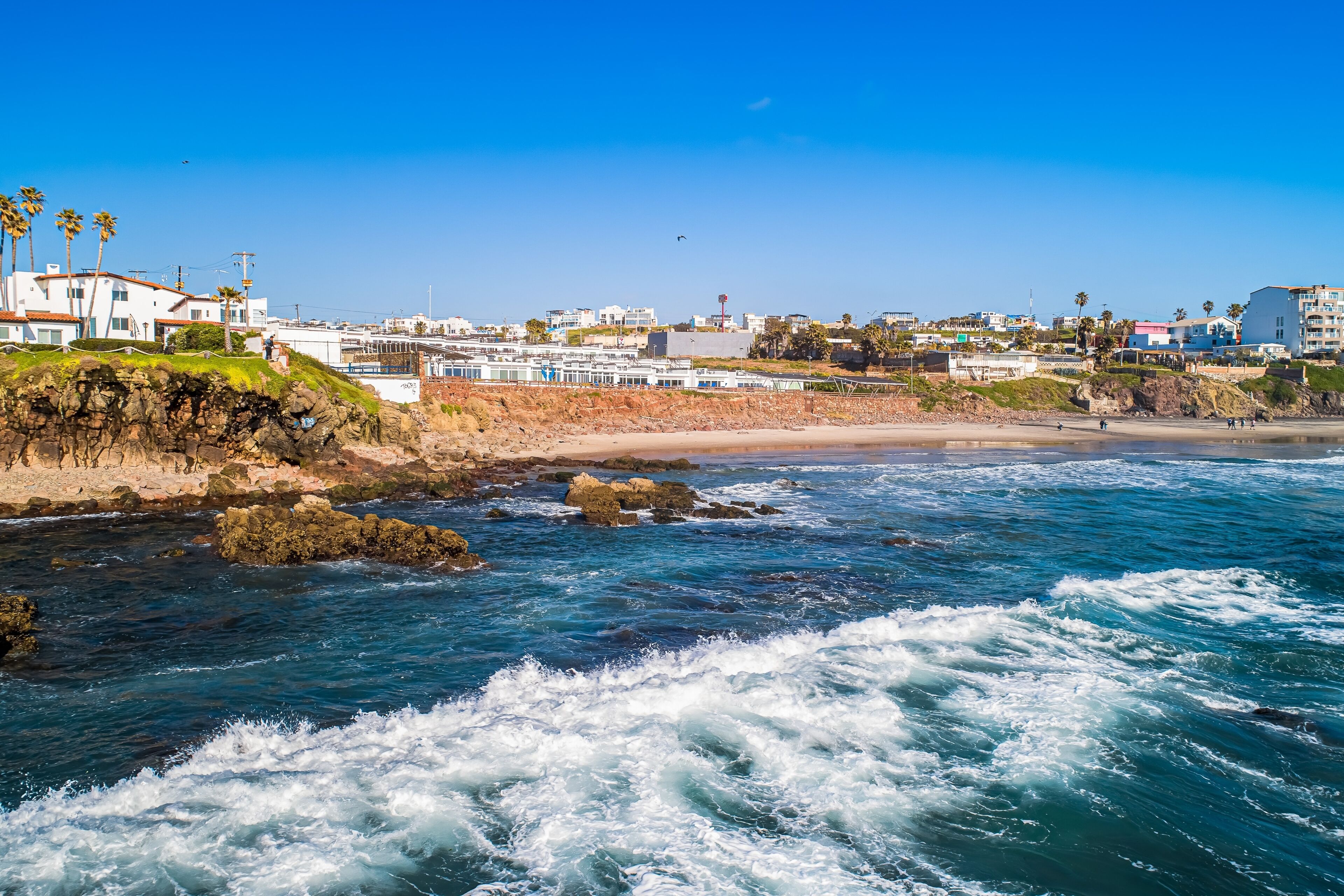 Zona costera con costa rocosa, olas y edificios al fondo.