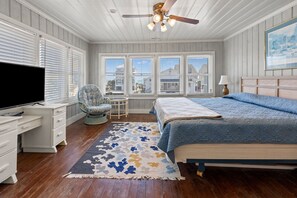 Ein Schlafzimmer mit Bett, Fernseher, Sessel, Deckenventilator und großen Fenstern.