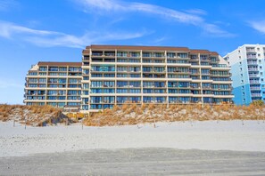 Ein mehrstöckiges Hotel mit Balkonen, ein sandiger Strand im Vordergrund und strahlend blauer Himmel.