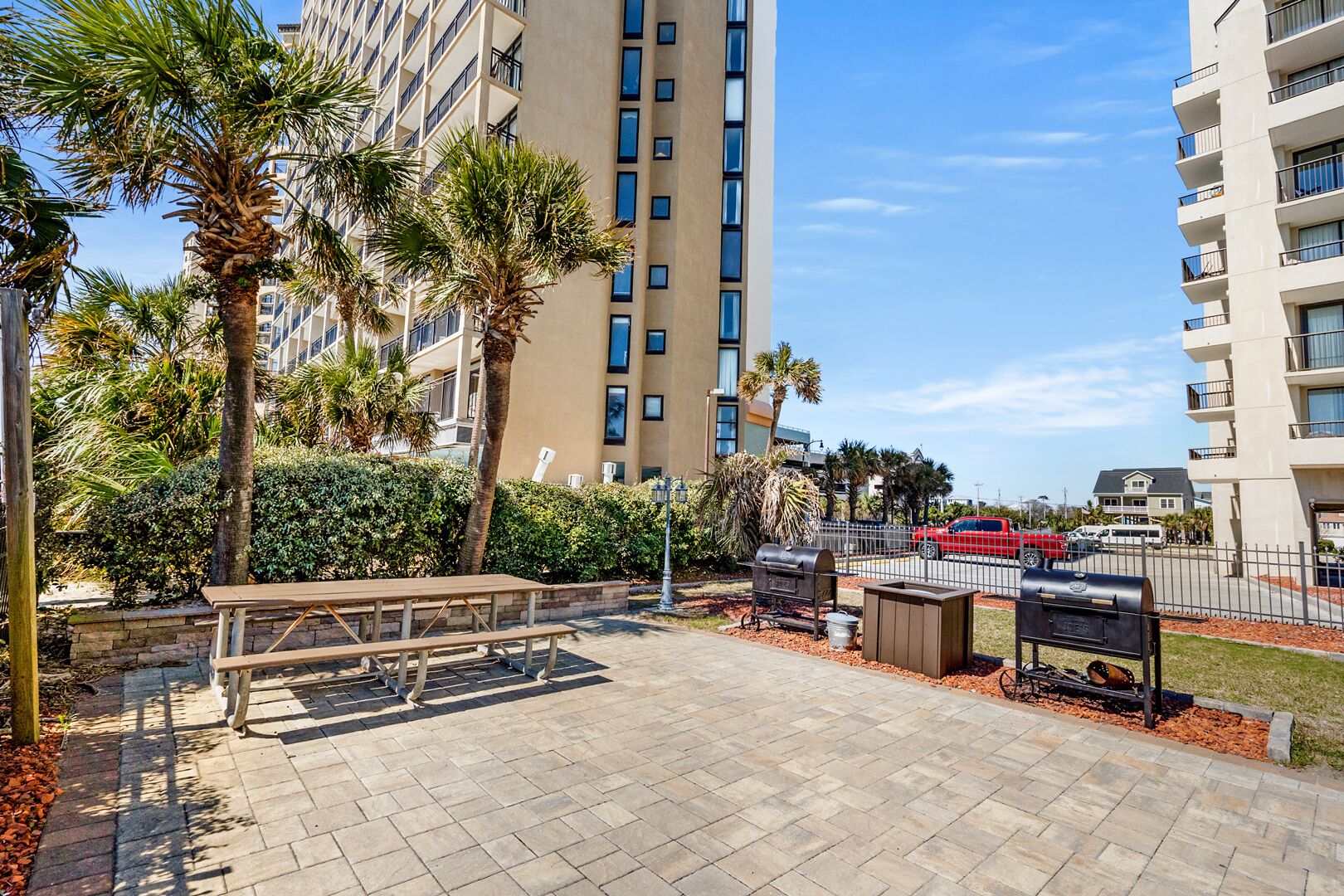 A paved outdoor area with a picnic table, barbecue grill, and trash bins.