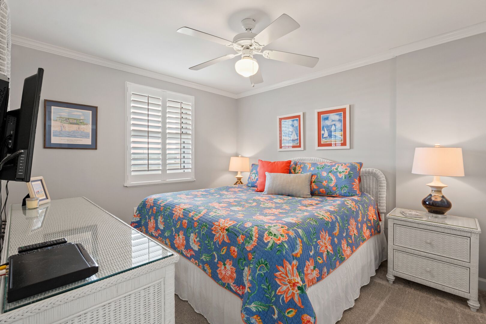 A bedroom with a bed, two bedside tables, a desk, a chair, a ceiling fan, and a window with blinds.