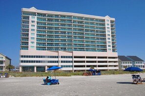 A multi-story hotel with a large parking structure, outdoor seating, and a clear blue sky. - Oceanfront 3BR condo with indoor pool, lazy river, hot tub, and private balcony in Cherry Grove (North Myrtle Beach)