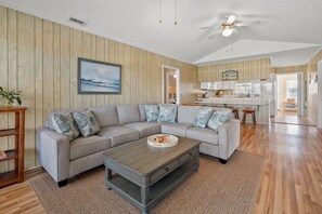 Ein Wohnzimmer mit einer grauen Eckcouch, einem Couchtisch und Holzboden. Im Hintergrund befindet sich eine Küche mit weißen Schränken und einem Essbereich.