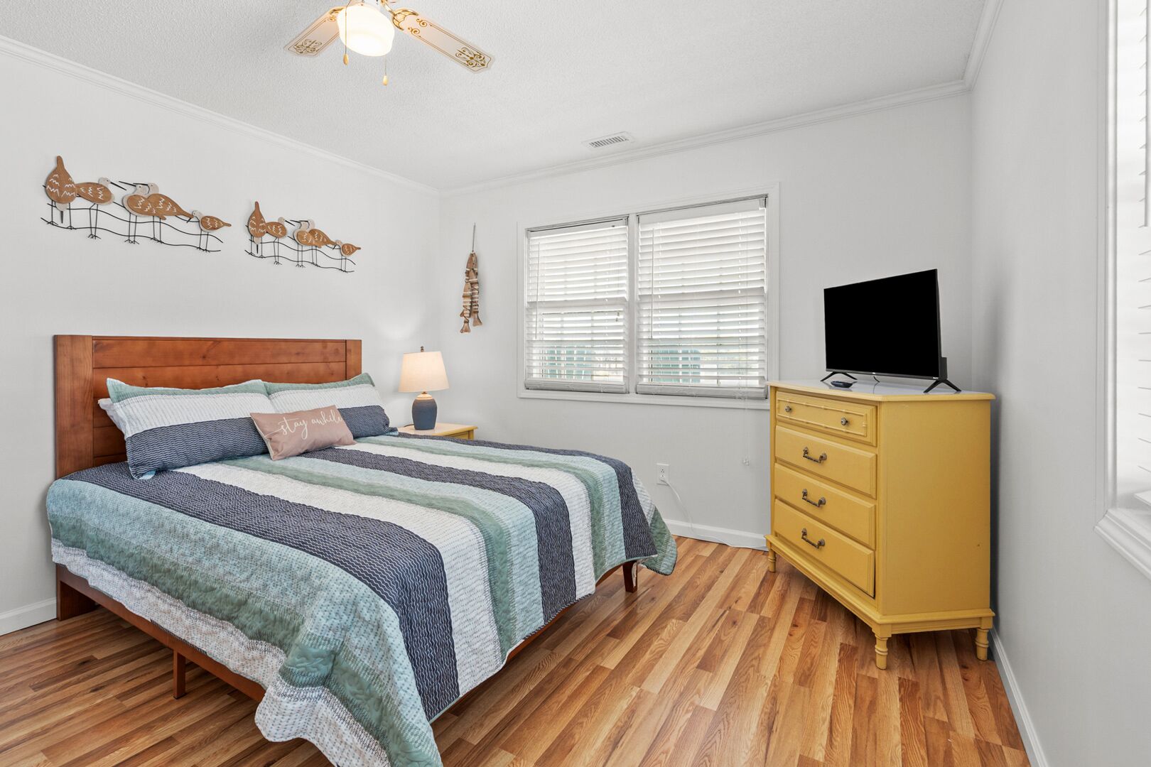 Quarto com cama de madeira, mesa de cabeceira, luminária, cômoda, televisão e janela com persianas.
