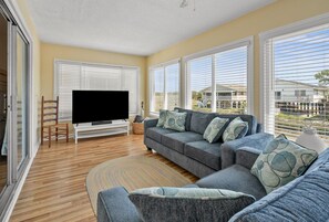 Ein Wohnzimmer mit Fernseher, Sofa und großen Fenstern.
