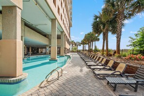 A swimming pool with lounge chairs, palm trees, and a building with a tiled roof. - Bahama Sands 901- a luxury ocean view condo in North Myrtle Beach (North Myrtle Beach)