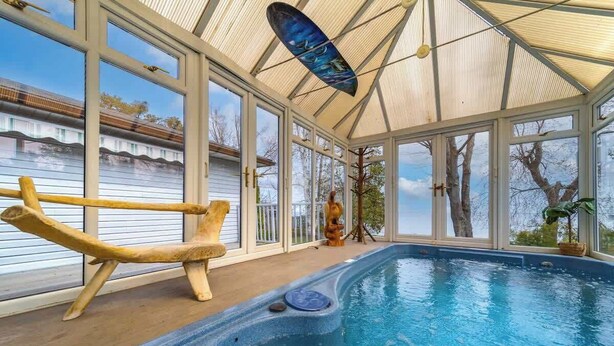 Une piscine intérieure dot ée d’un toit en verre, d’une terrasse en bois et d’un jacuzzi.