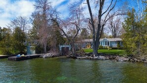 Une maison au bord d’un lac, avec un quai, un petit abri de jardin et un arbre au premier plan.