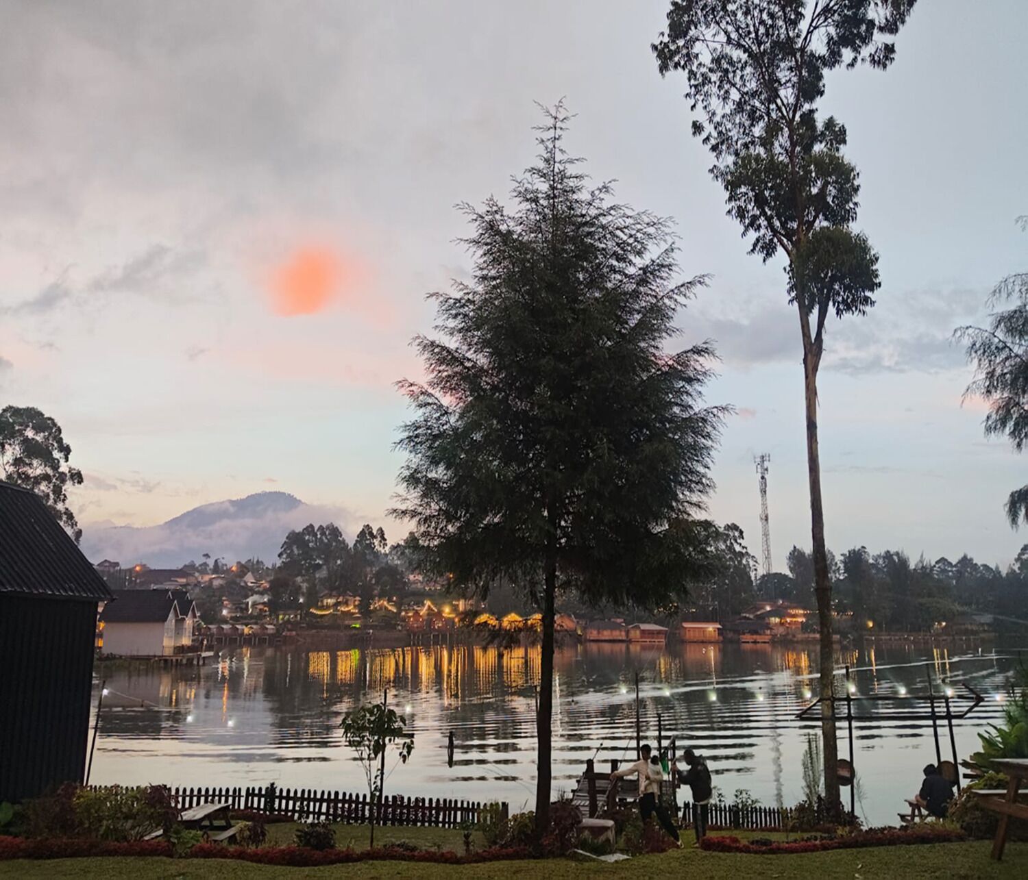 Vista al lago con valla de madera, árboles y edificios al fondo.