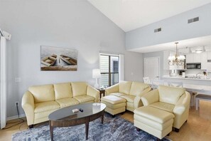 Un salon avec un ensemble de meubles de couleur beige, une table basse et un tableau représentant des bateaux accroché au mur.