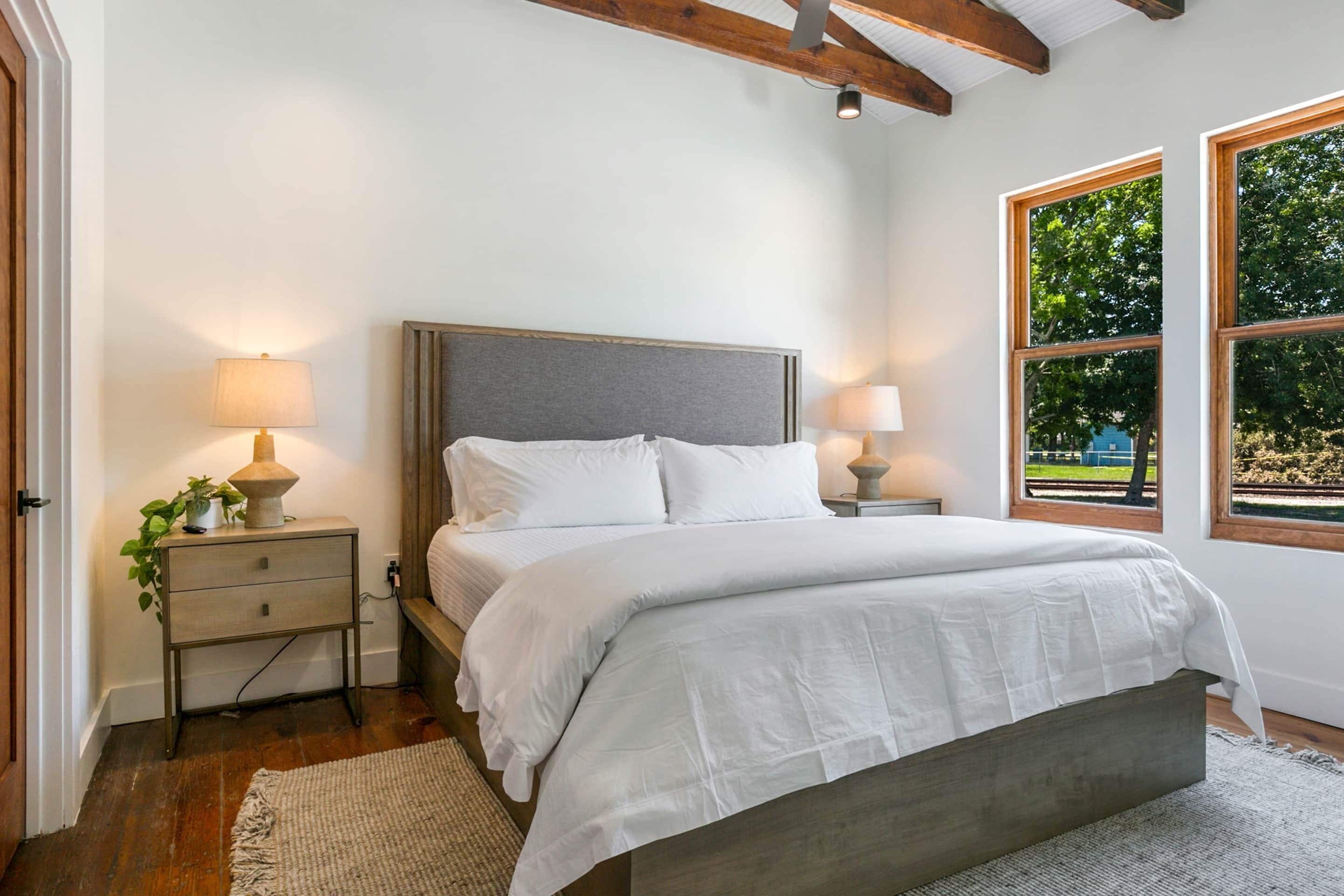 A bedroom with a bed, bedside tables, lamps, a plant, and a view through large windows.