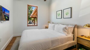 A bedroom with a large bed, a window with a view, framed artwork, and a nightstand with a lamp. - Commander 6 Farragut Lofts, Old Town, City Park (Bay Saint Louis)