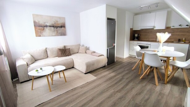 A modern living room with a sectional sofa, a dining area with a round table and chairs, and a kitchen with white cabinets and a refrigerator. - Beautiful large apartment with super roof terrace (Walsrode)