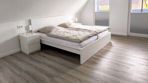 A bedroom with a white bed, a nightstand, a lamp, and a window with a view of buildings. - Beautiful large apartment with super roof terrace (Walsrode)