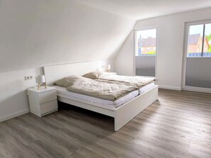 A bedroom with a white bed, a nightstand, a lamp, and a window with a view of buildings. - Beautiful large apartment with super roof terrace (Walsrode)