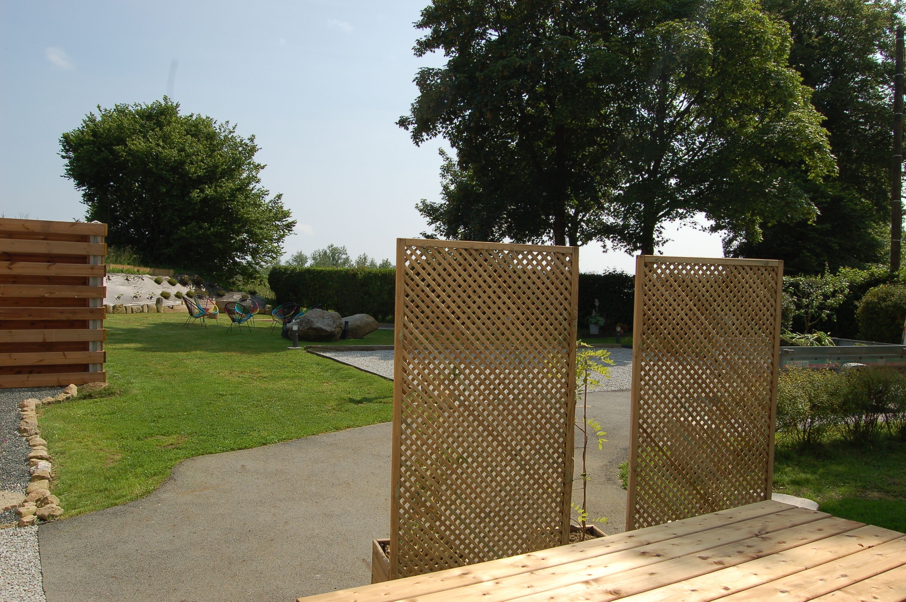 Une terrasse en bois agrémentée d’un treillis décoratif, un chemin pavé et une zone herbeuse avec des arbres et des rochers.