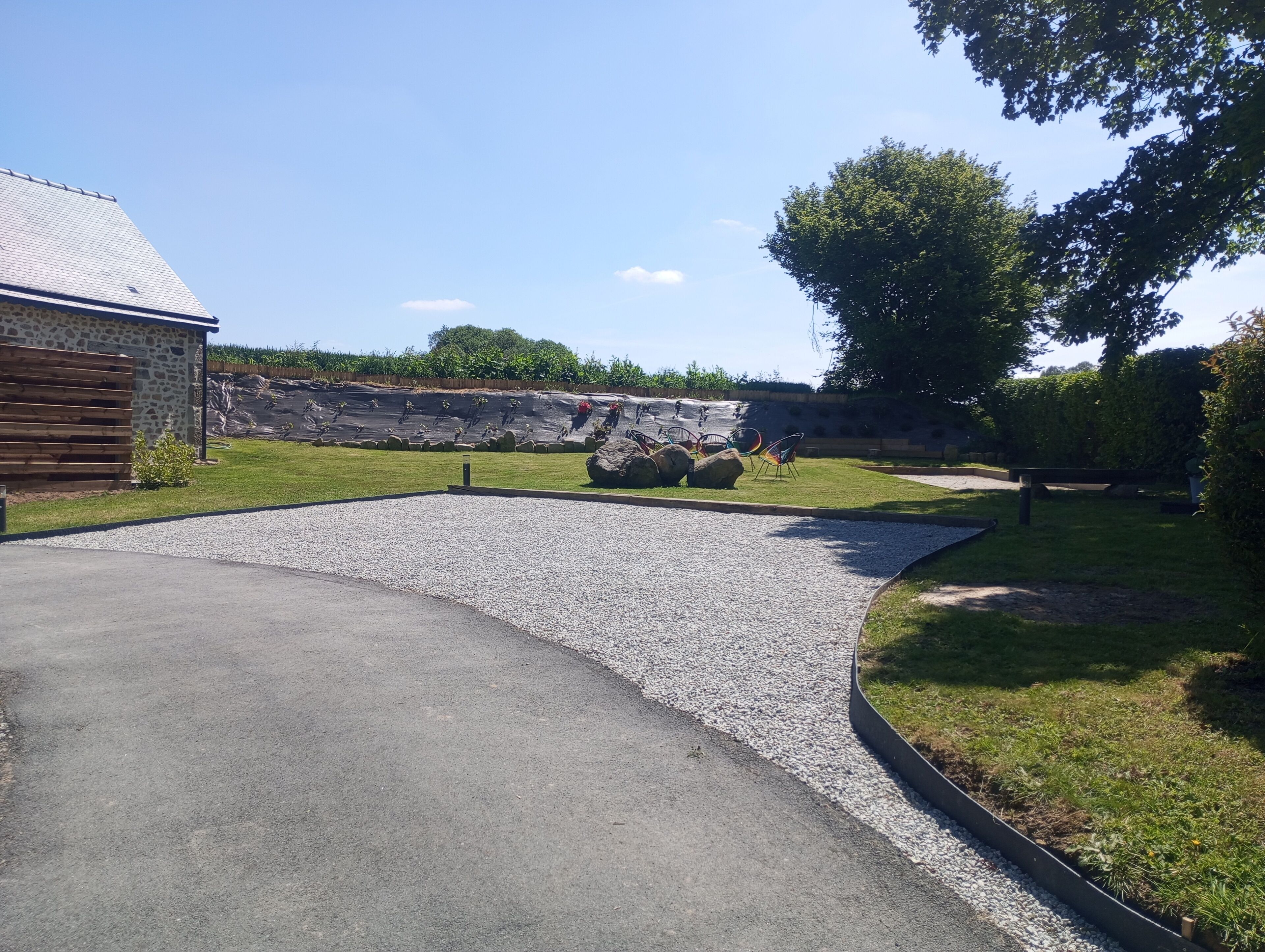 A paved driveway leading to a building with a stone facade, a grassy area with a rock feature, and a tree in the background.