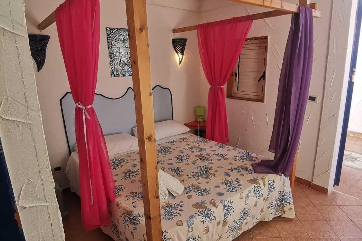 A room with a four-poster bed, a window with shutters, a wall-mounted light fixture, and a tiled floor.