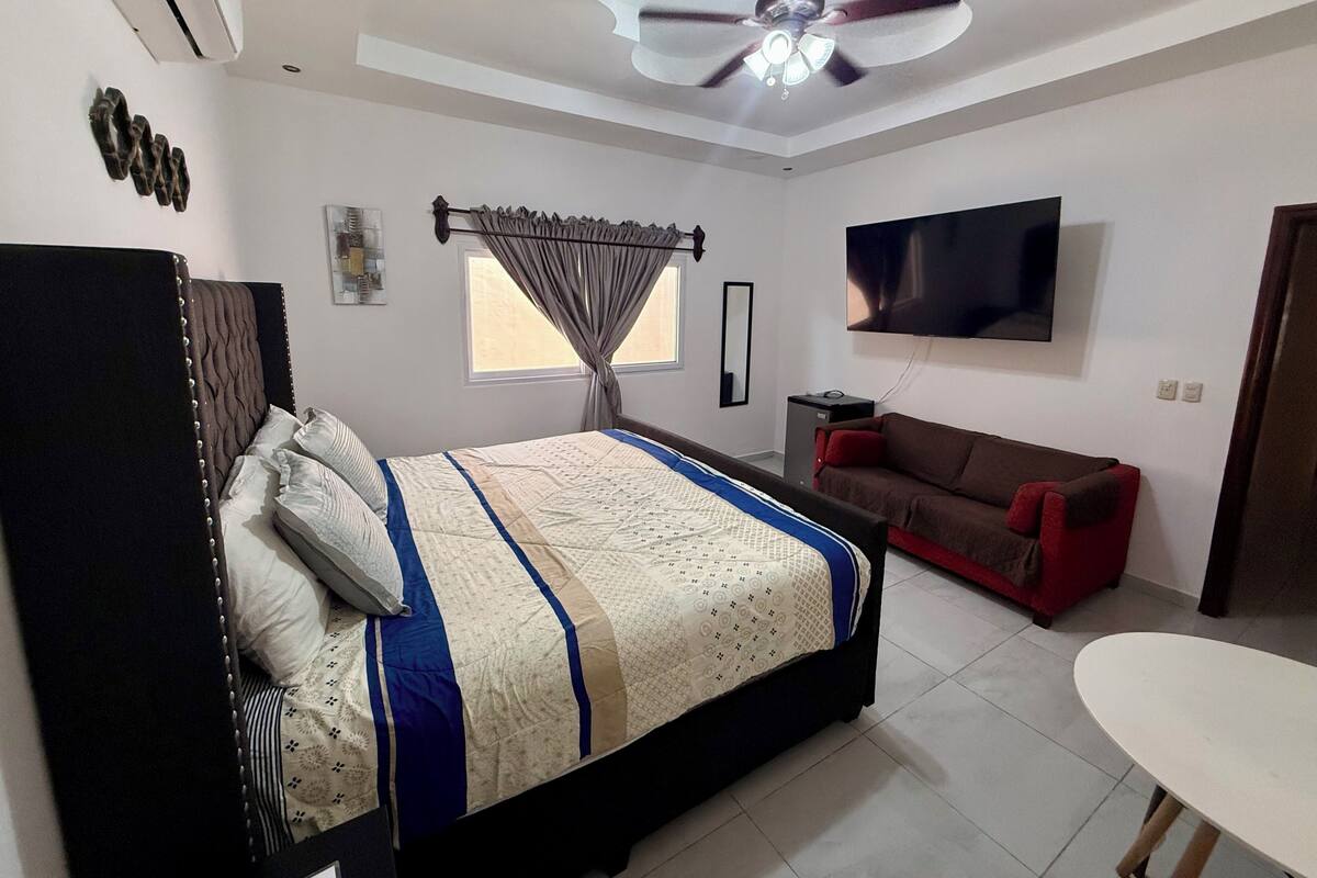Un dormitorio con cama, ventilador de techo, televisión, una mesita y una ventana con cortinas.