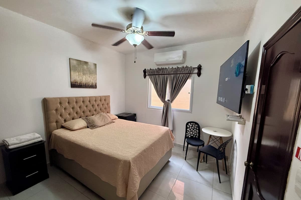 Un dormitorio con cama, ventilador de techo, televisión, escritorio con silla y ventana con cortinas.