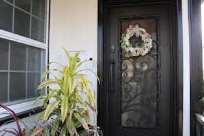 Une porte noire ornée d’une guirlande décorative, un pot de fleurs et une fenêtre sur le côté gauche.