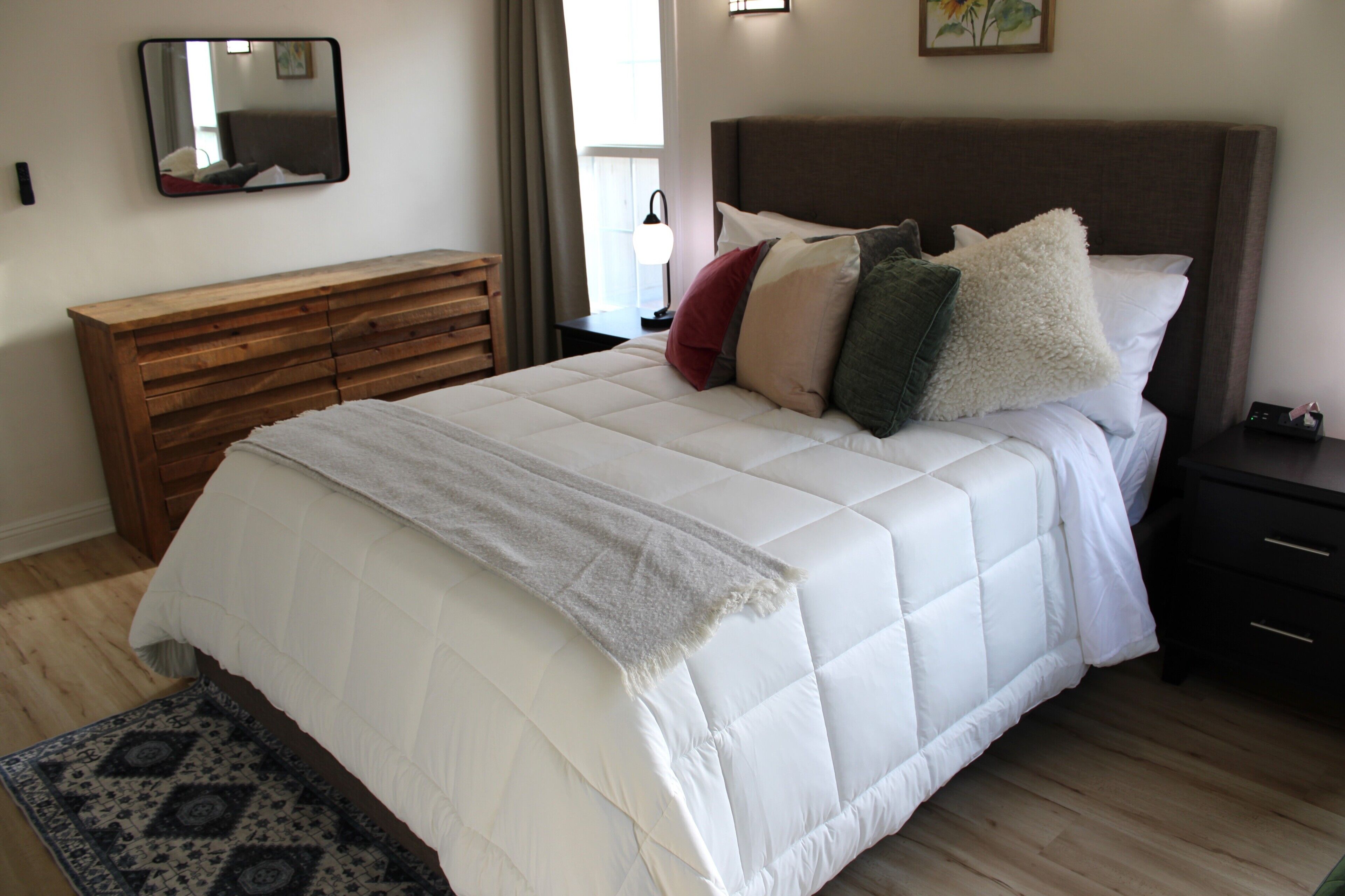 A bedroom with a bed, a wooden dresser, a nightstand, a lamp, and a mirror on the wall.