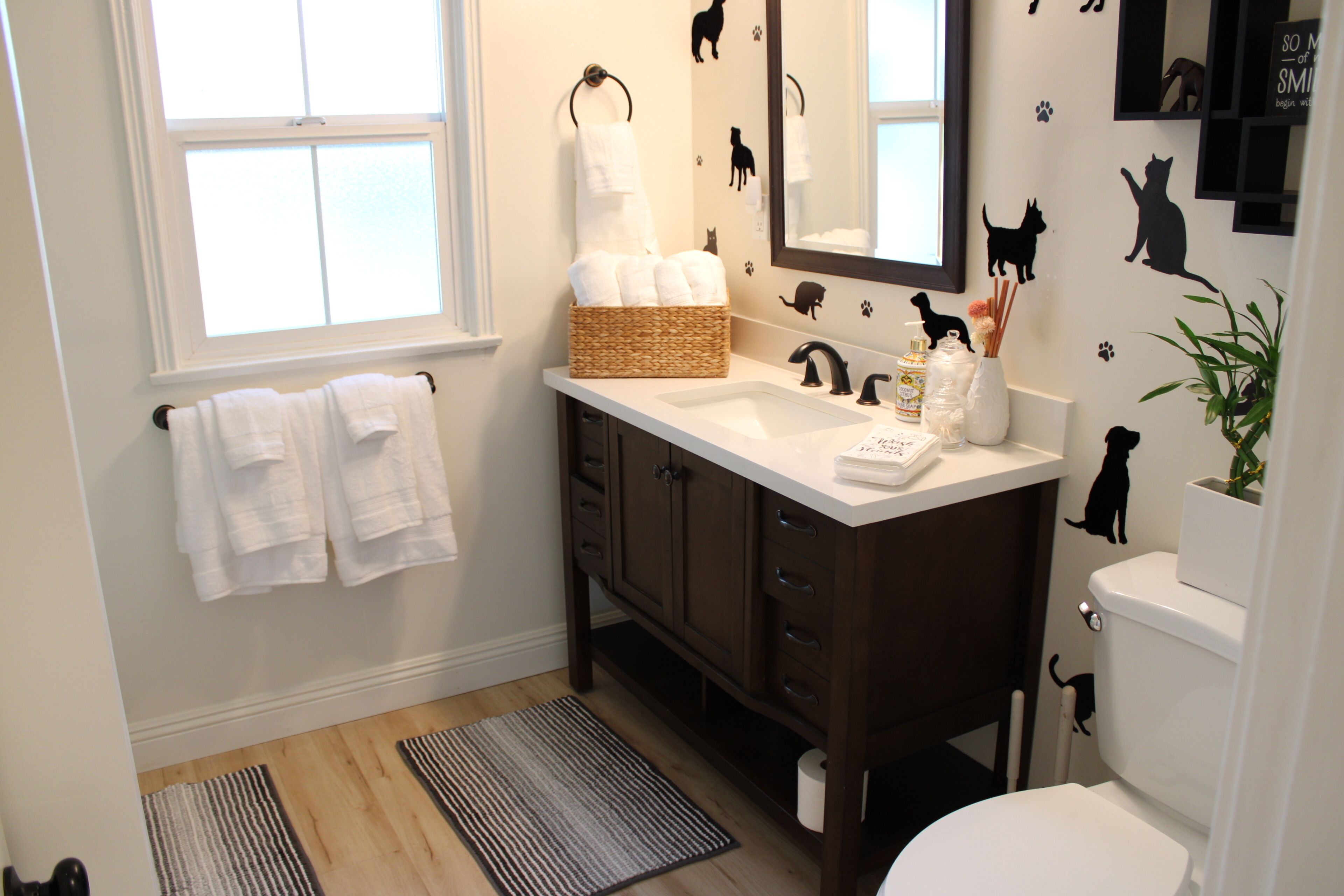 Un bagno con un lavandino bianco, un mobile bagno in legno scuro, un cesto di asciugamani bianchi, uno specchio e decorazioni murali con gatti e cani.