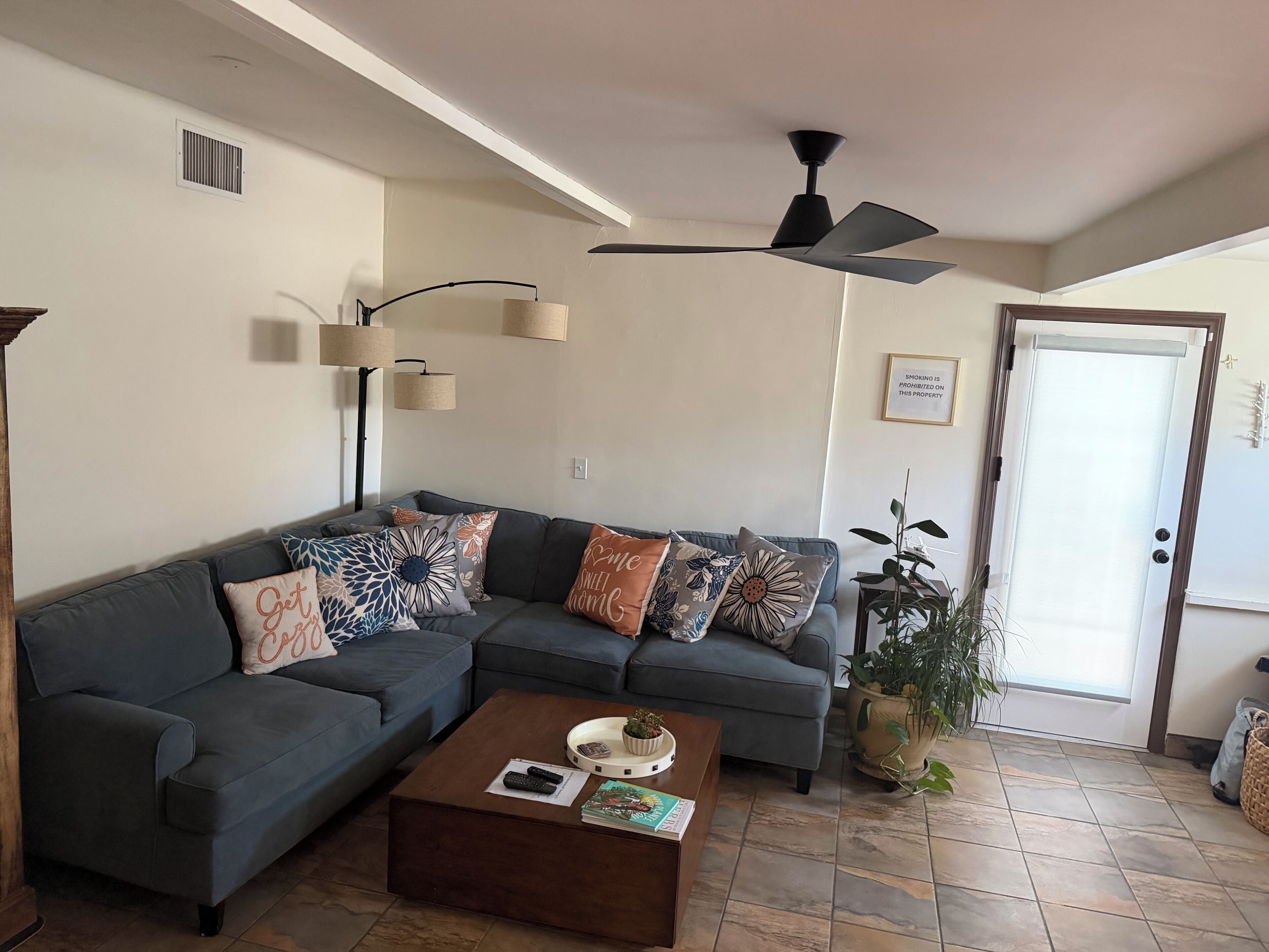 A living room with a sectional sofa, a coffee table, a ceiling fan, and a door with a sign.