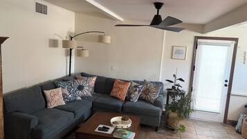 A living room with a sectional sofa, a coffee table, a ceiling fan, and a door with a sign.