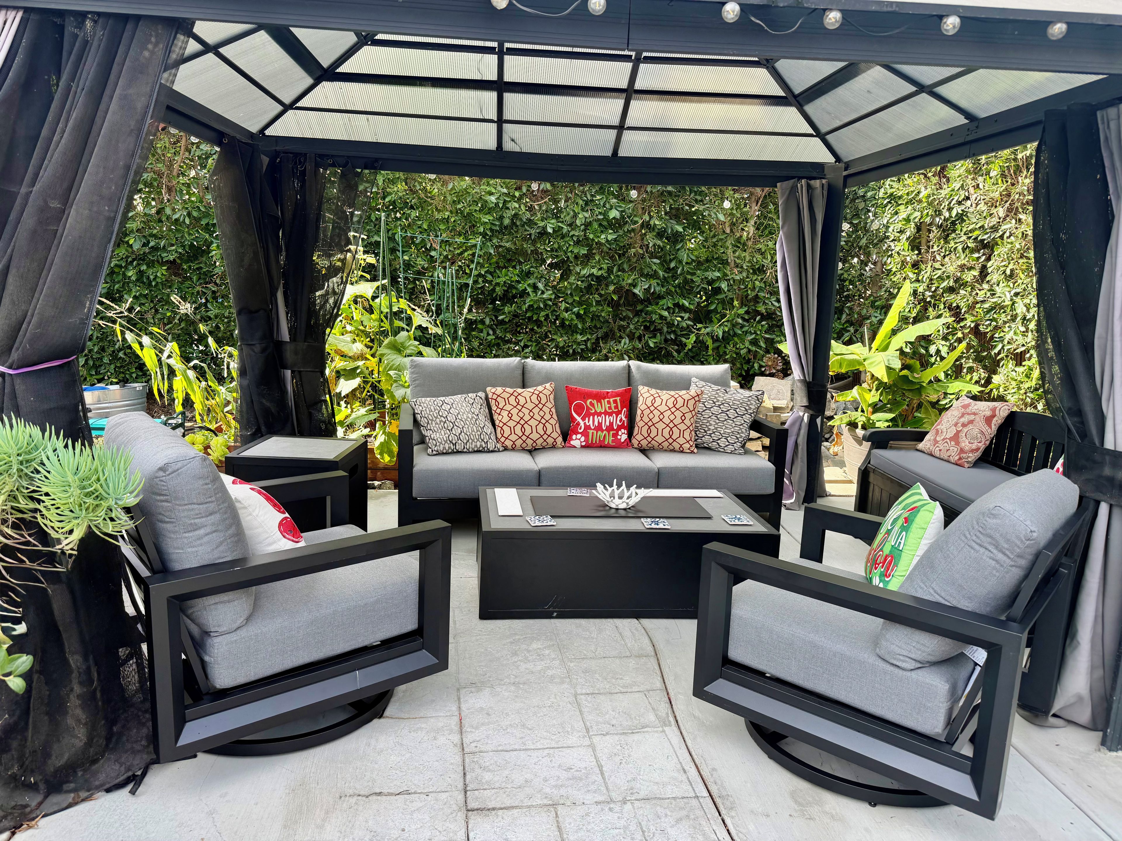 A covered outdoor seating area with a sofa and chairs, surrounded by greenery.