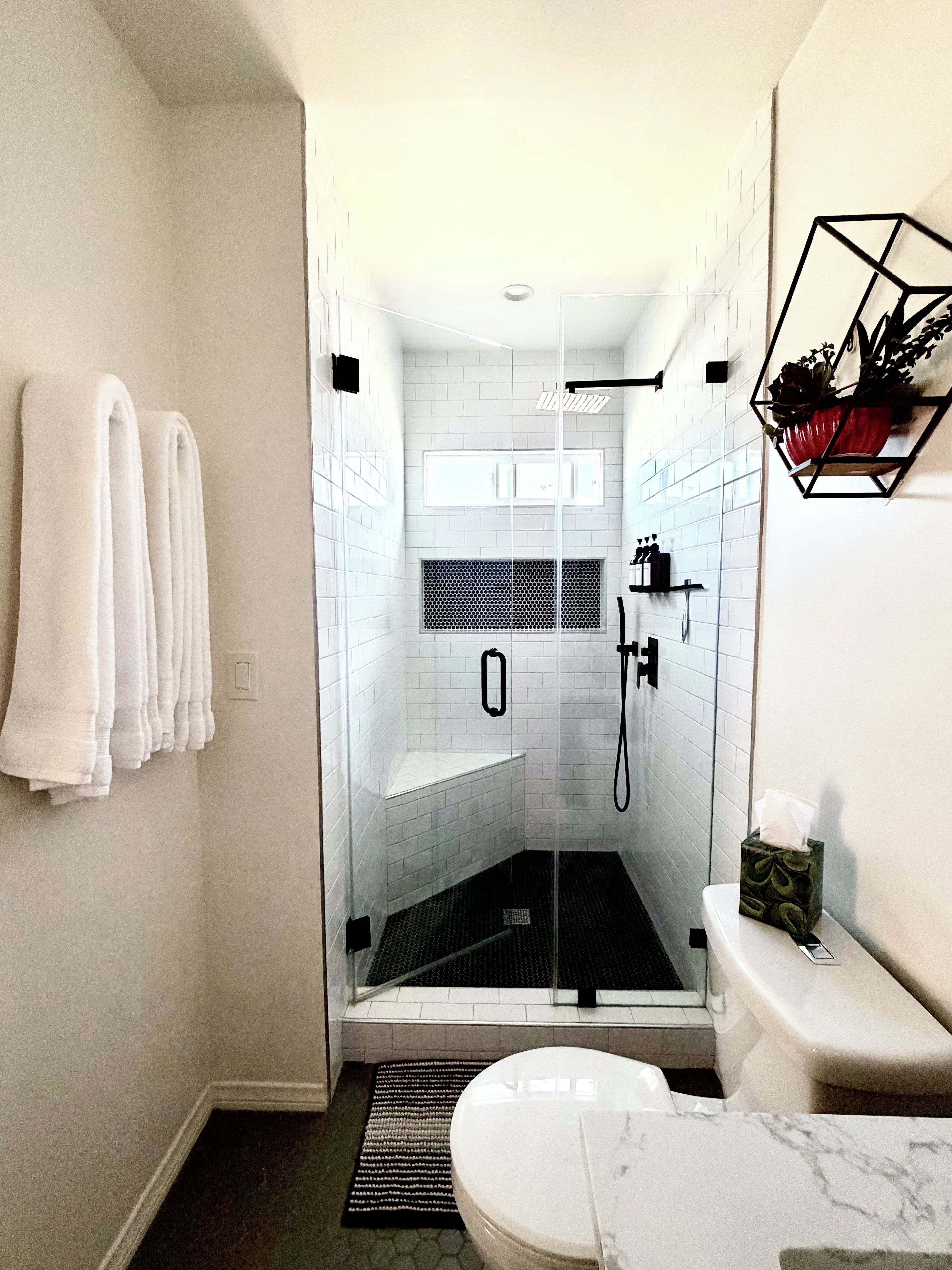 Une salle de bain moderne avec une douche à parois de verre, des murs recouverts de carreaux blancs, des toilettes et un porte-serviettes.
